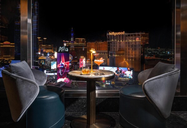 Un coin bar avec deux fauteuils en cuir et une table ronde, offrant une vue sur le paysage urbain, avec des bâtiments illuminés en arrière-plan.