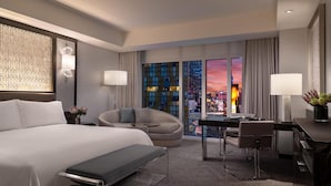A modern hotel room with a large bed, a desk, a chair, and a view of the cityscape at sunset.