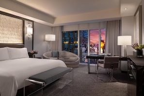 Een moderne hotelkamer met een groot bed, een bureau, een stoel en uitzicht op de stad bij zonsondergang.