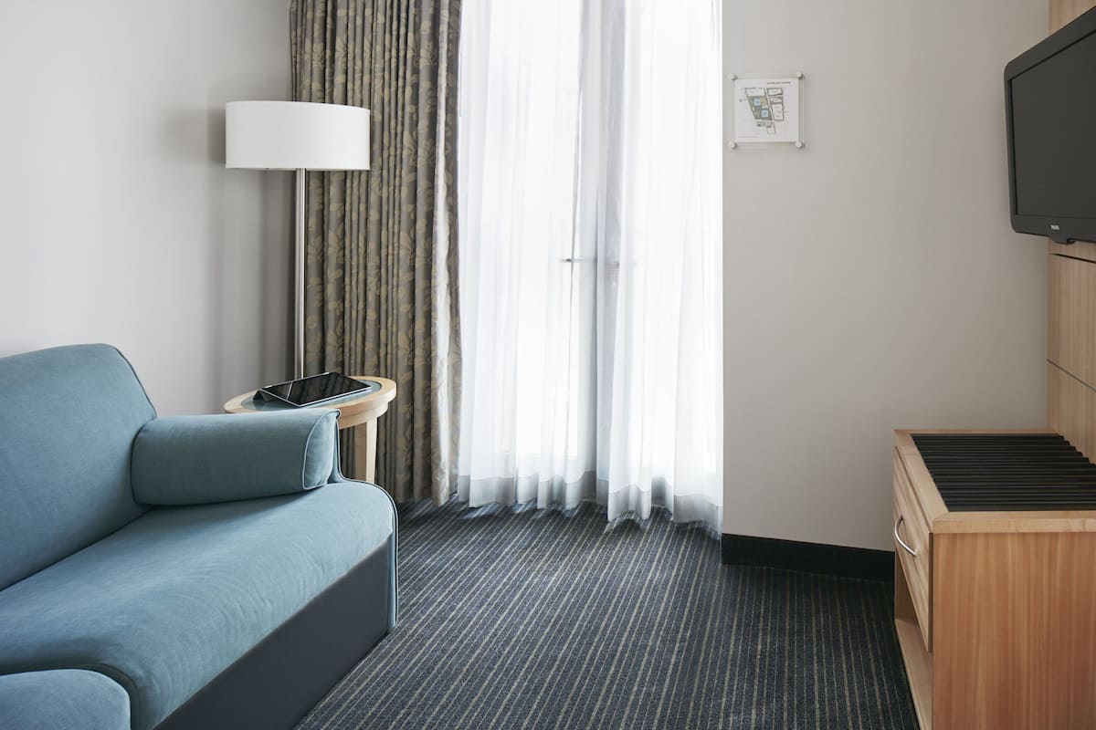 A hotel room with a sofa, a lamp, a TV, a wooden cabinet, and a window with curtains.