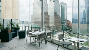 A modern indoor balcony with a city view, featuring a table and chairs, potted plants, and a glass railing. - World Center Hotel (New York)