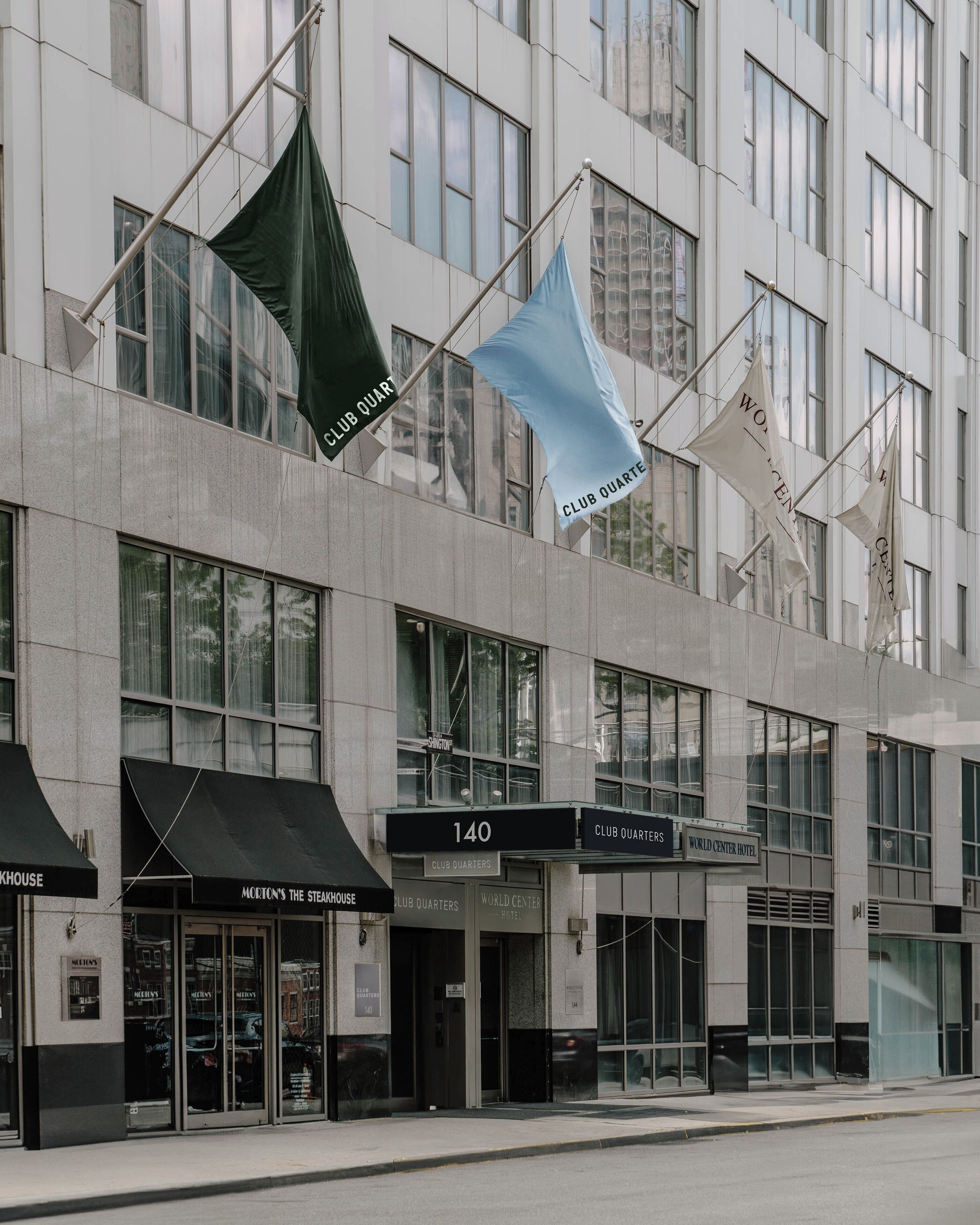 A modern building with multiple flags, including 'Club Quarters' and 'World Center Hotel' signs, and storefronts like 'Morton's The Steakhouse'.