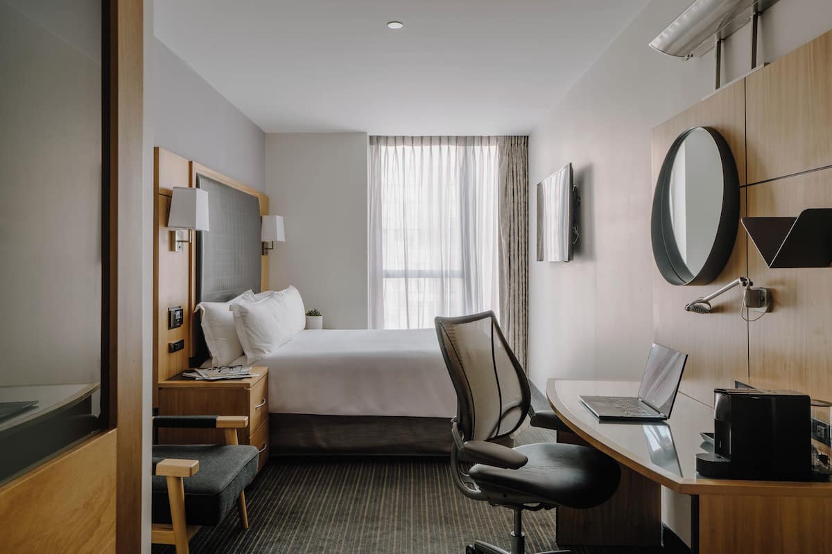 A hotel room with a bed, a desk with a laptop, a chair, a desk lamp, a flat-screen TV, a bedside table with a lamp, and a window with curtains.