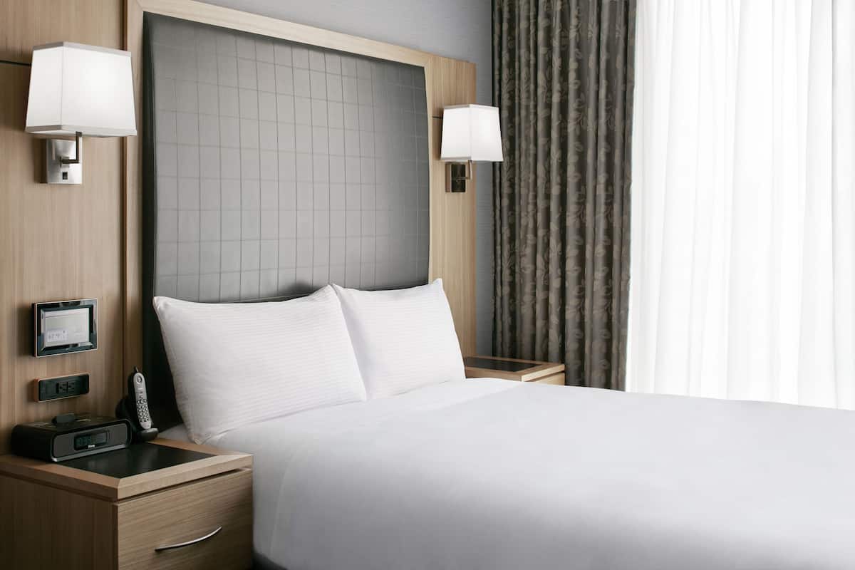 A hotel room with a large bed, two bedside tables, a digital clock, a thermostat, and a telephone.