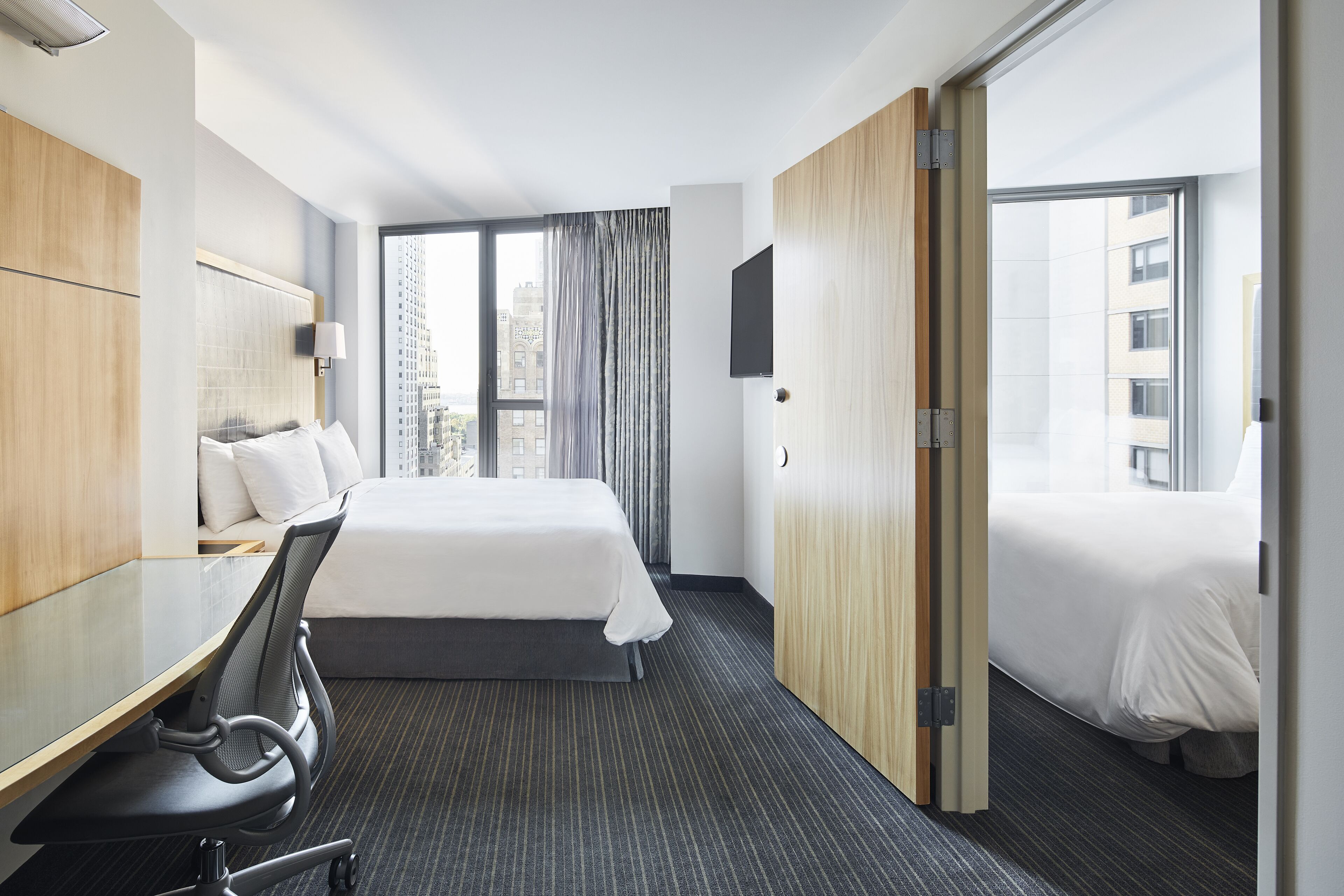 A hotel room with a large bed, a desk with a chair, a TV mounted on the wall, and a window with curtains.