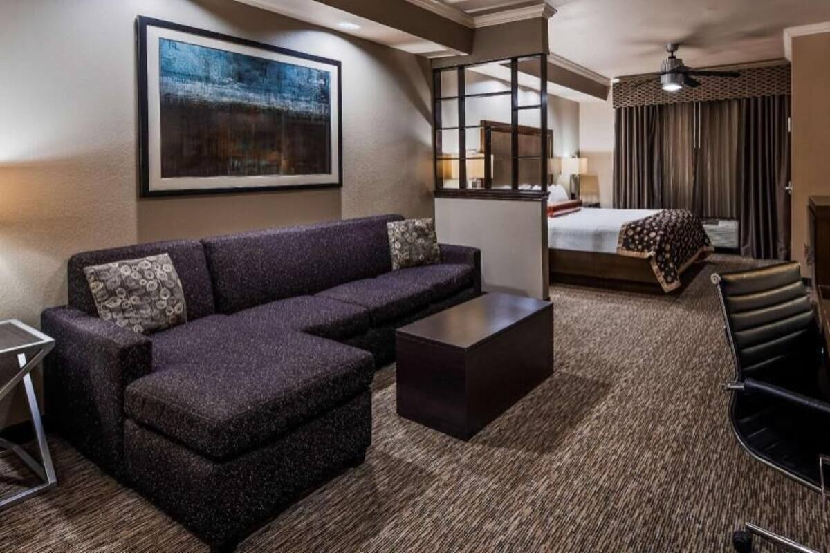 A hotel room with a sectional sofa, a small table, a bed with a patterned bedspread, a TV, and a large window.