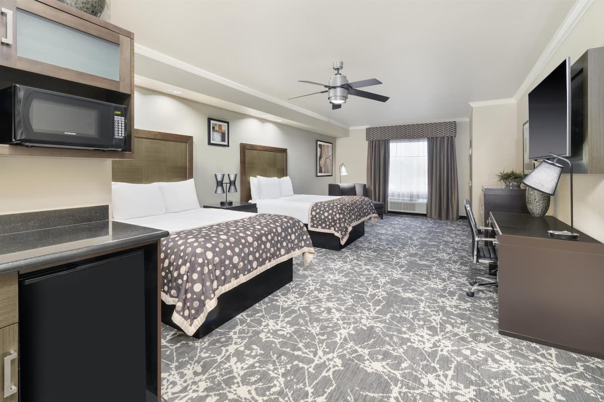 A hotel room with two beds, a desk, a microwave, a ceiling fan, and a large window with curtains.