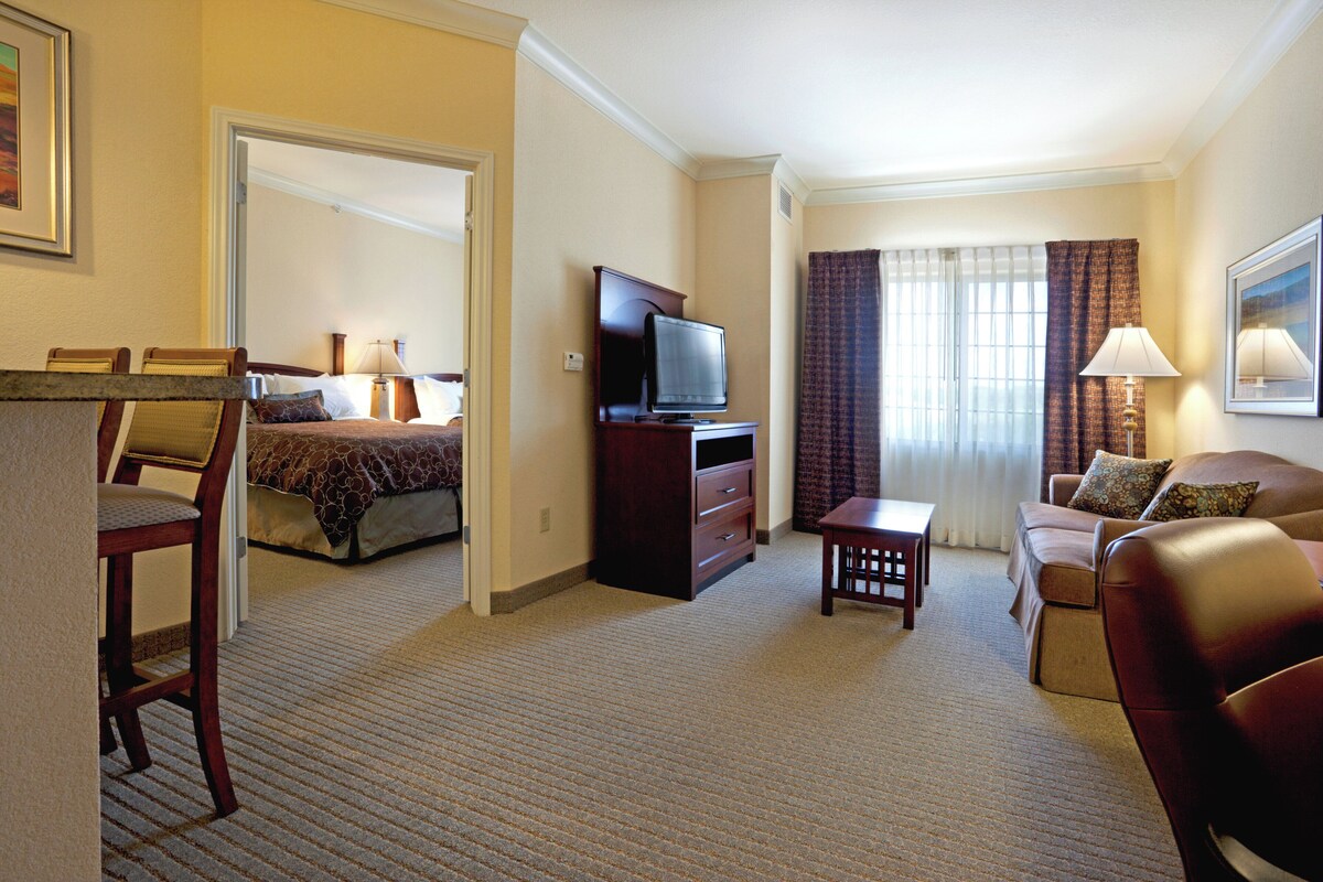 Habitación de hotel con cama, televisión, escritorio, silla, sofá y ventana con cortinas.