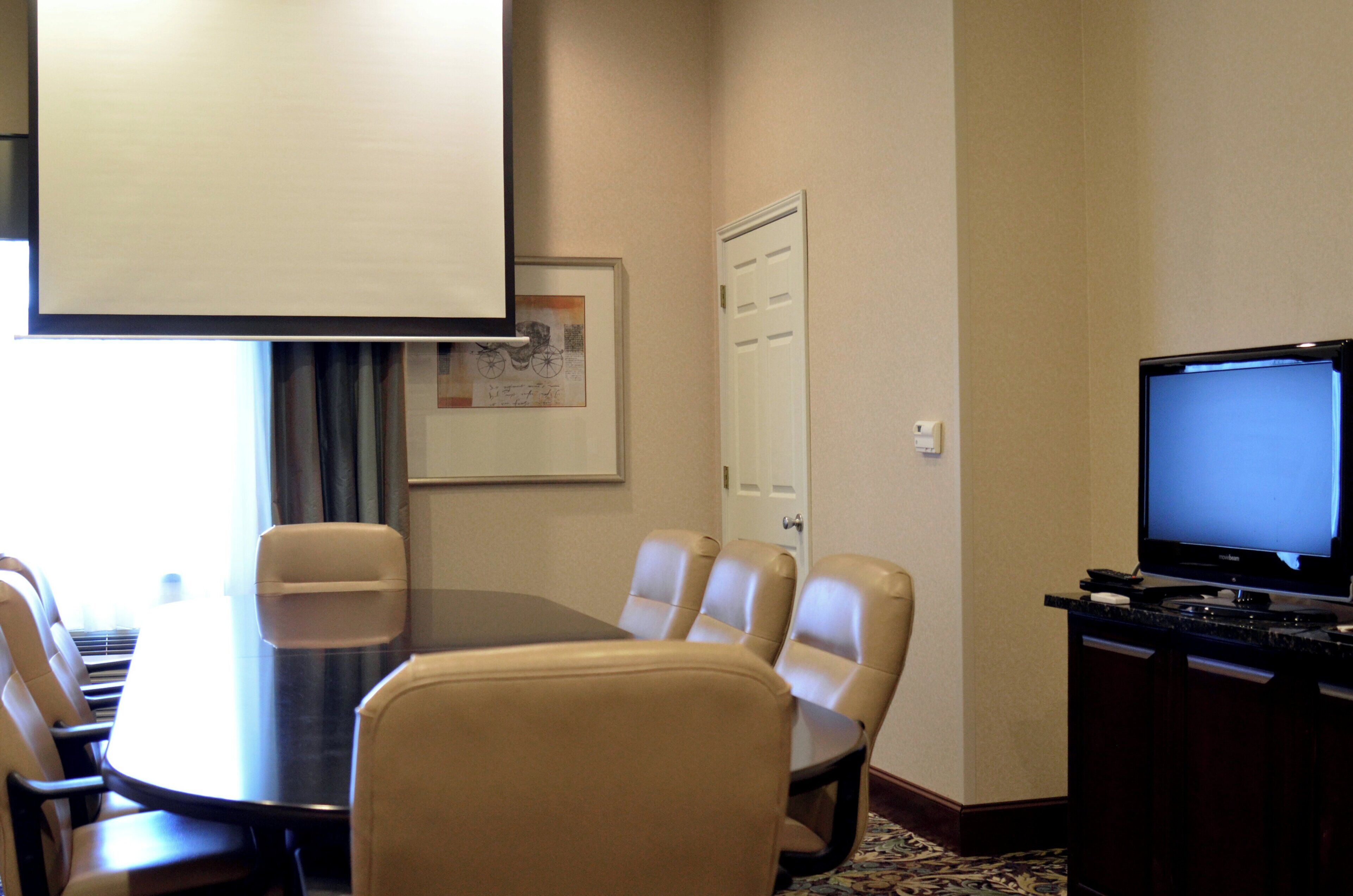 Un salón de conferencias con una mesa redonda, seis sillas, una pantalla de proyector, un televisor y una puerta.