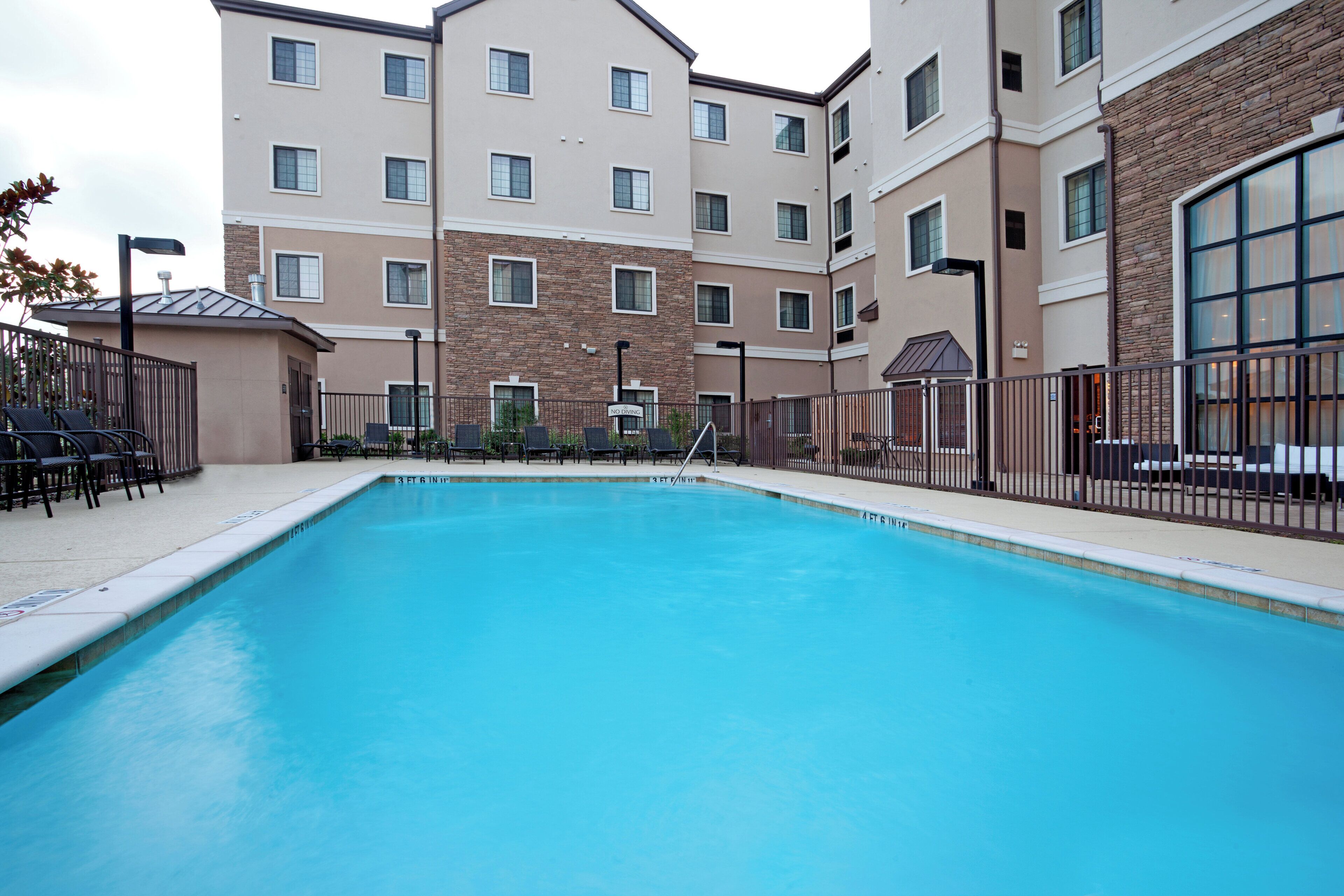 A swimming pool with a clear blue water surface, surrounded by a fence, and adjacent to a multi-story building with a brick facade and multiple windows.