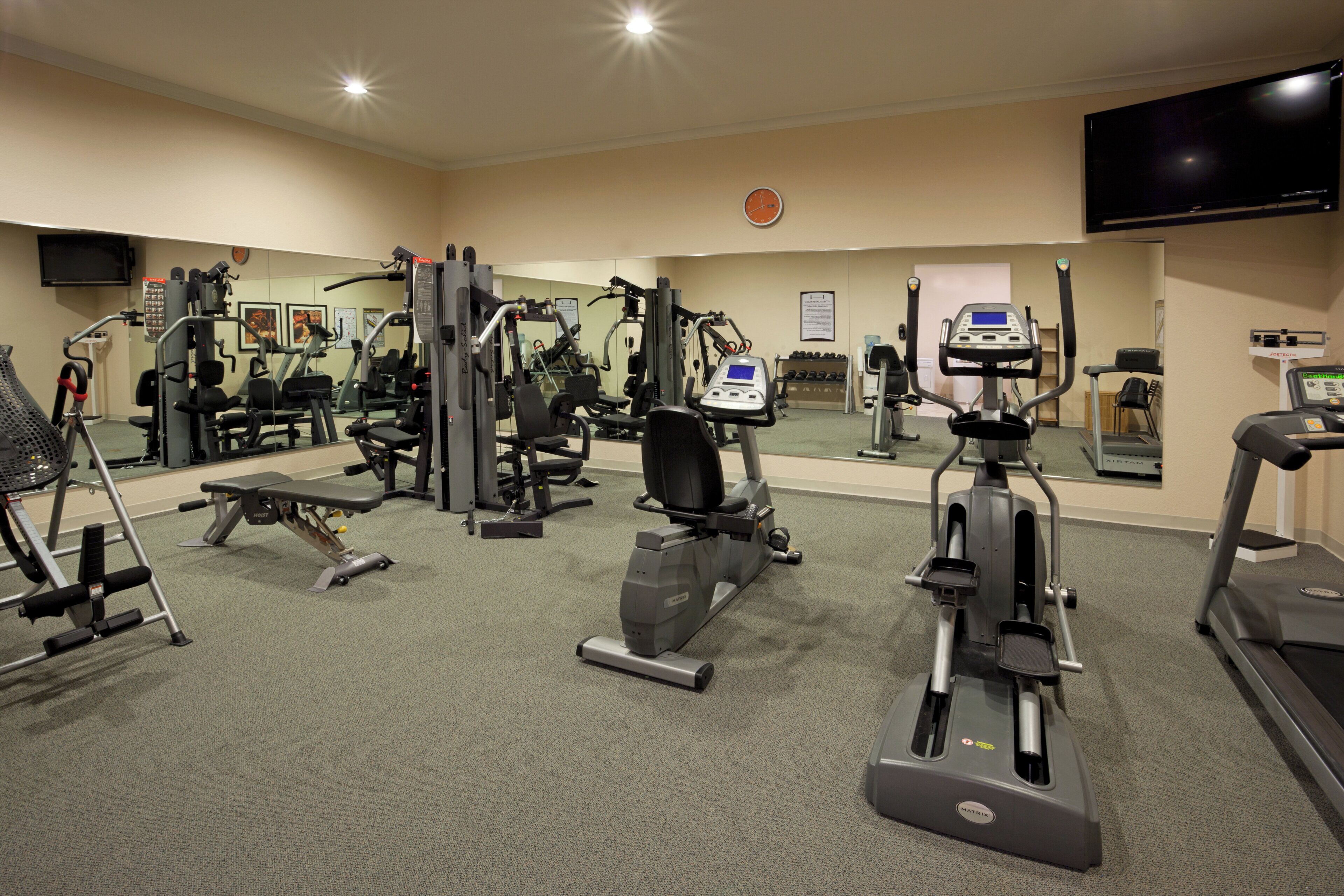 Un gimnasio con diferentes equipos de ejercicio, un televisor montado en la pared y un espejo que refleja el equipo.