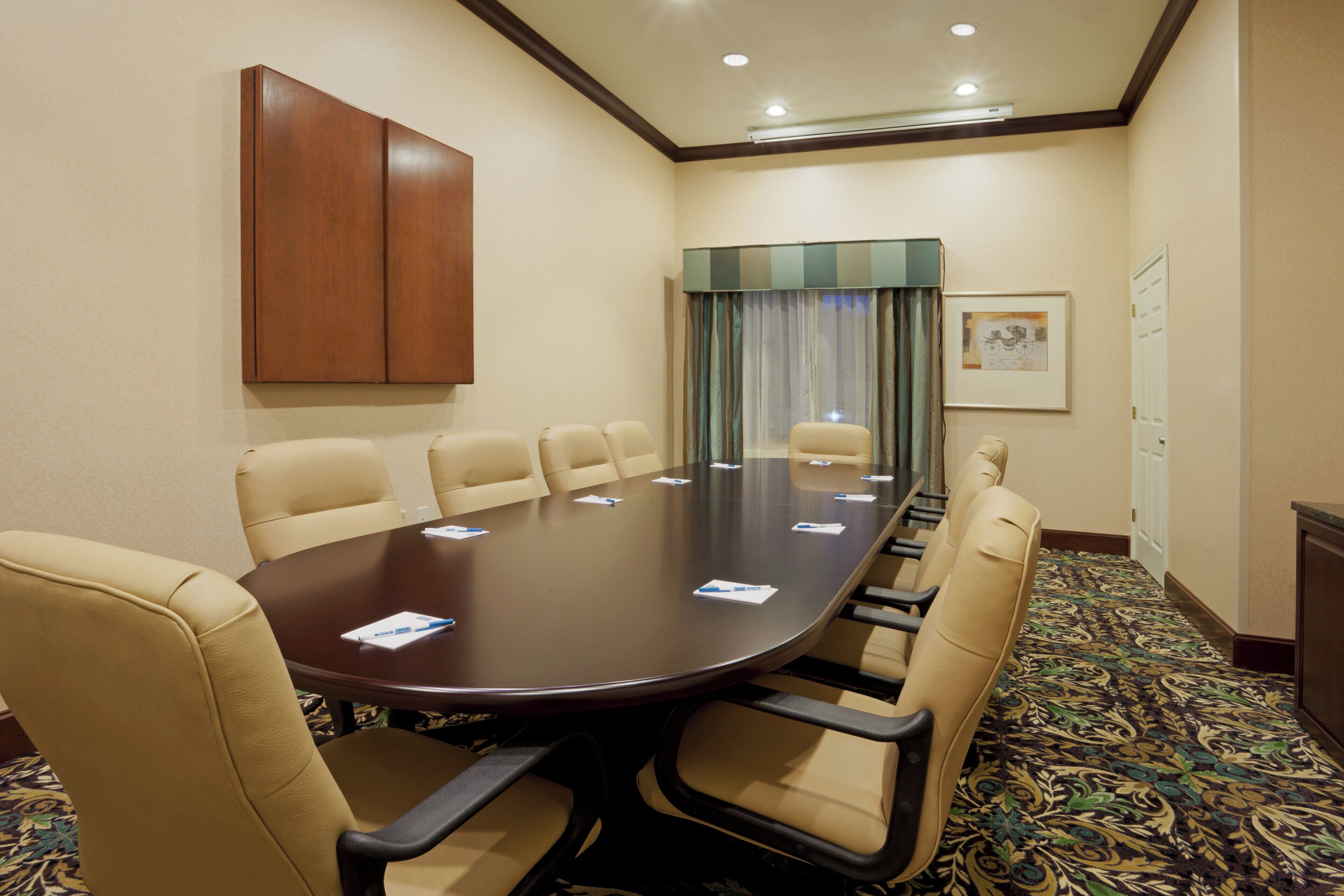 Un salón de conferencias con una gran mesa, seis sillas, un armario de madera, una ventana con cortinas y un cuadro en la pared.