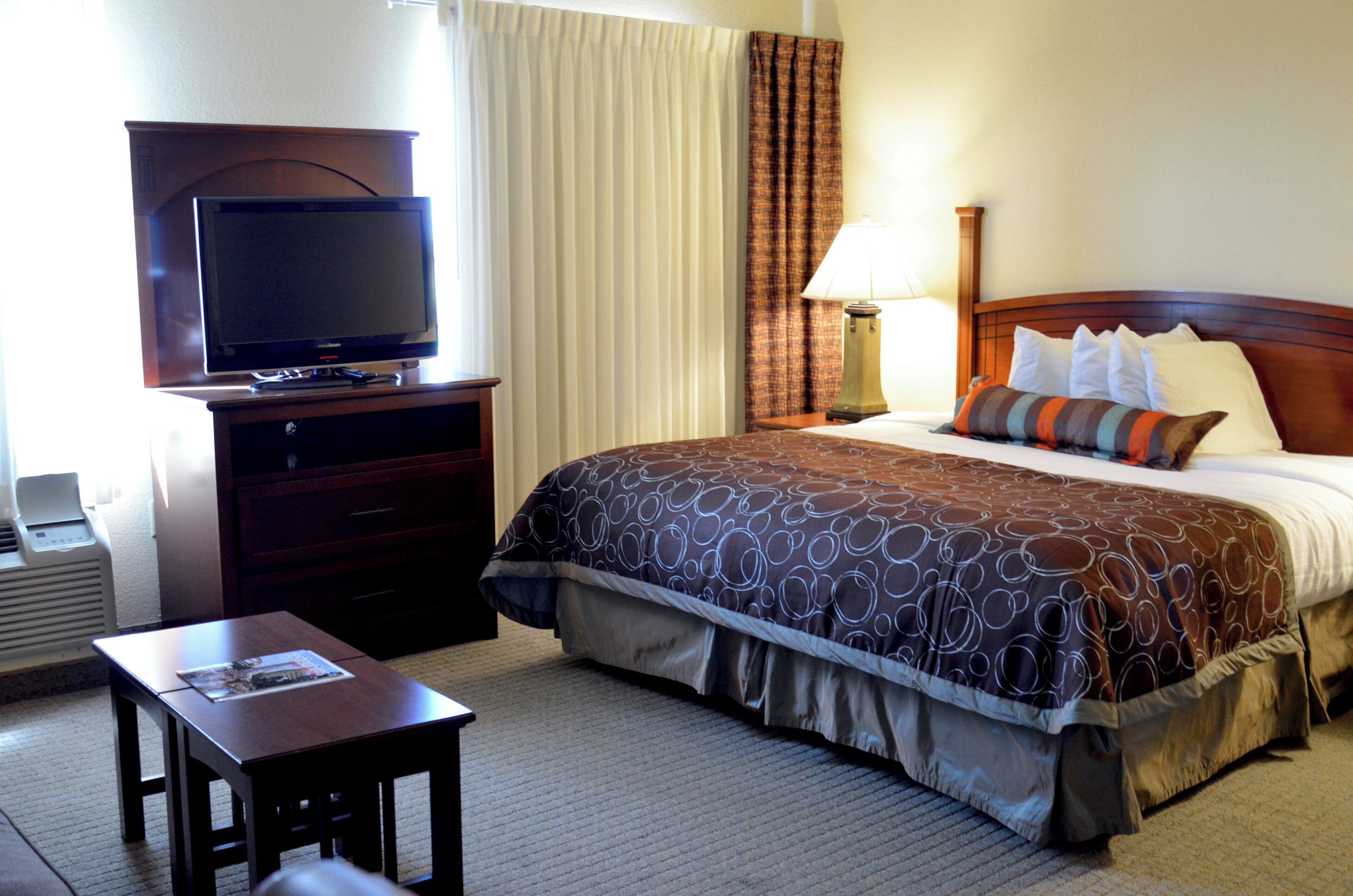 A hotel room with a bed, a television on a stand, a bedside table, and a lamp.