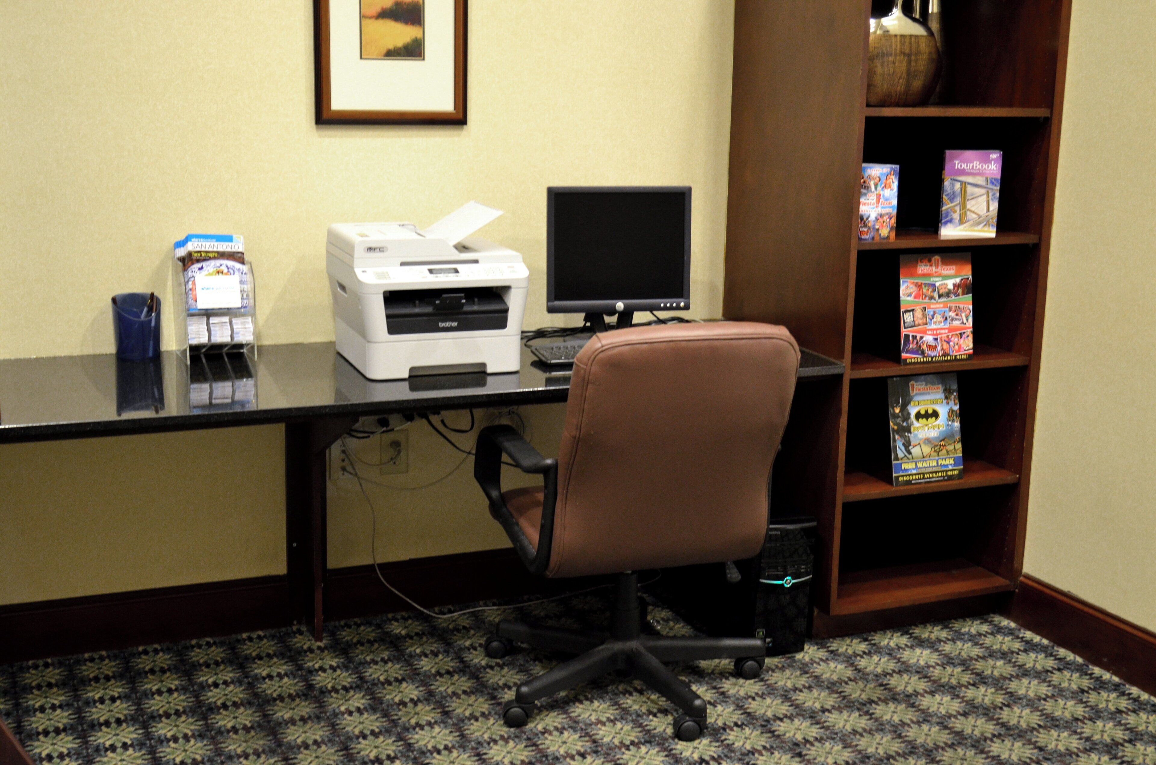 Habitación de hotel con escritorio, impresora, computadora, silla de oficina y una estantería con diversos objetos.