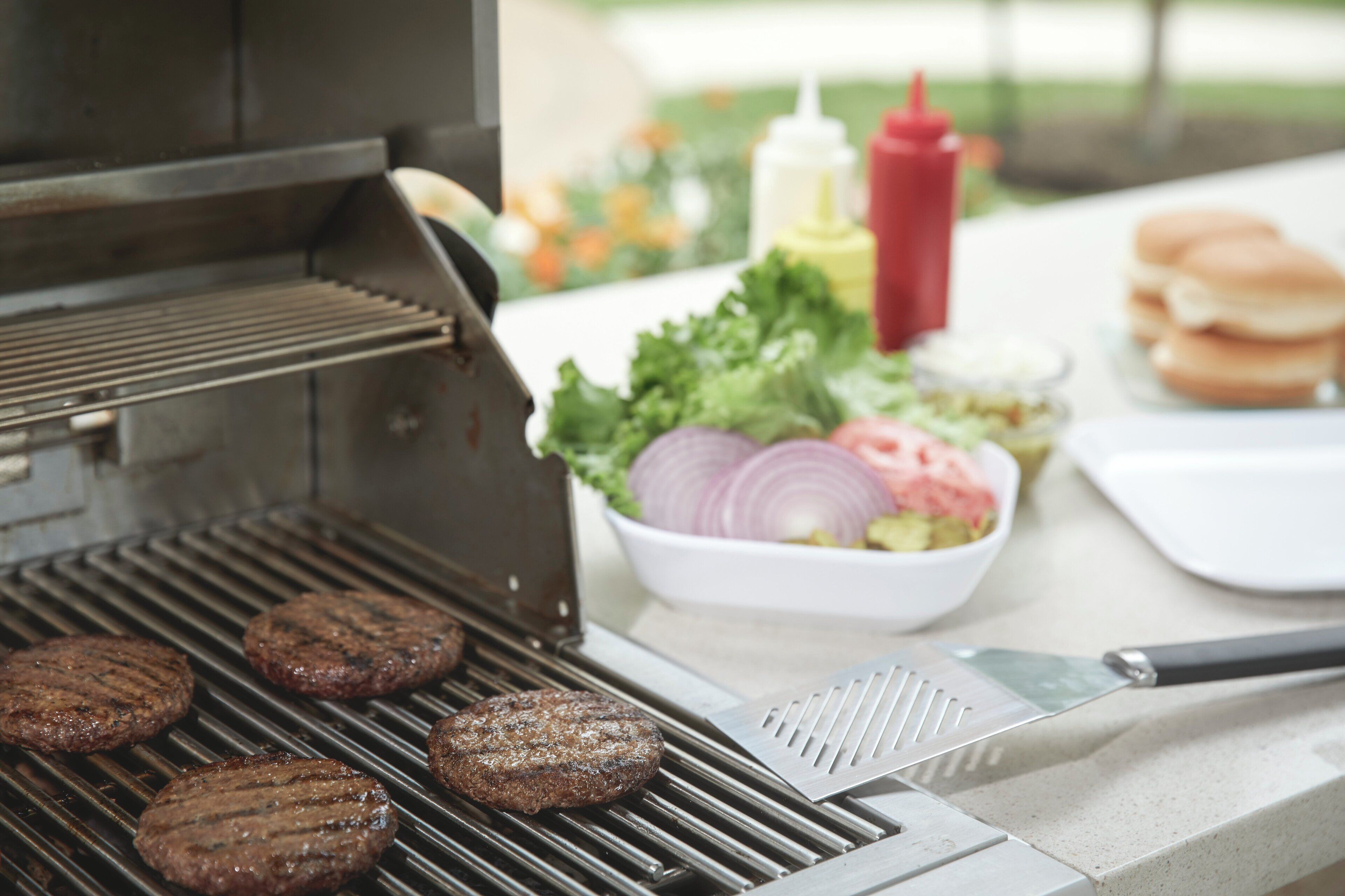 Un barbecue avec des hamburgers en train de cuire, une planche à découper avec un couteau et des condiments sur une table.