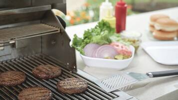 Un barbecue avec des hamburgers en train de cuire, une planche à découper avec un couteau et des condiments sur une table.
