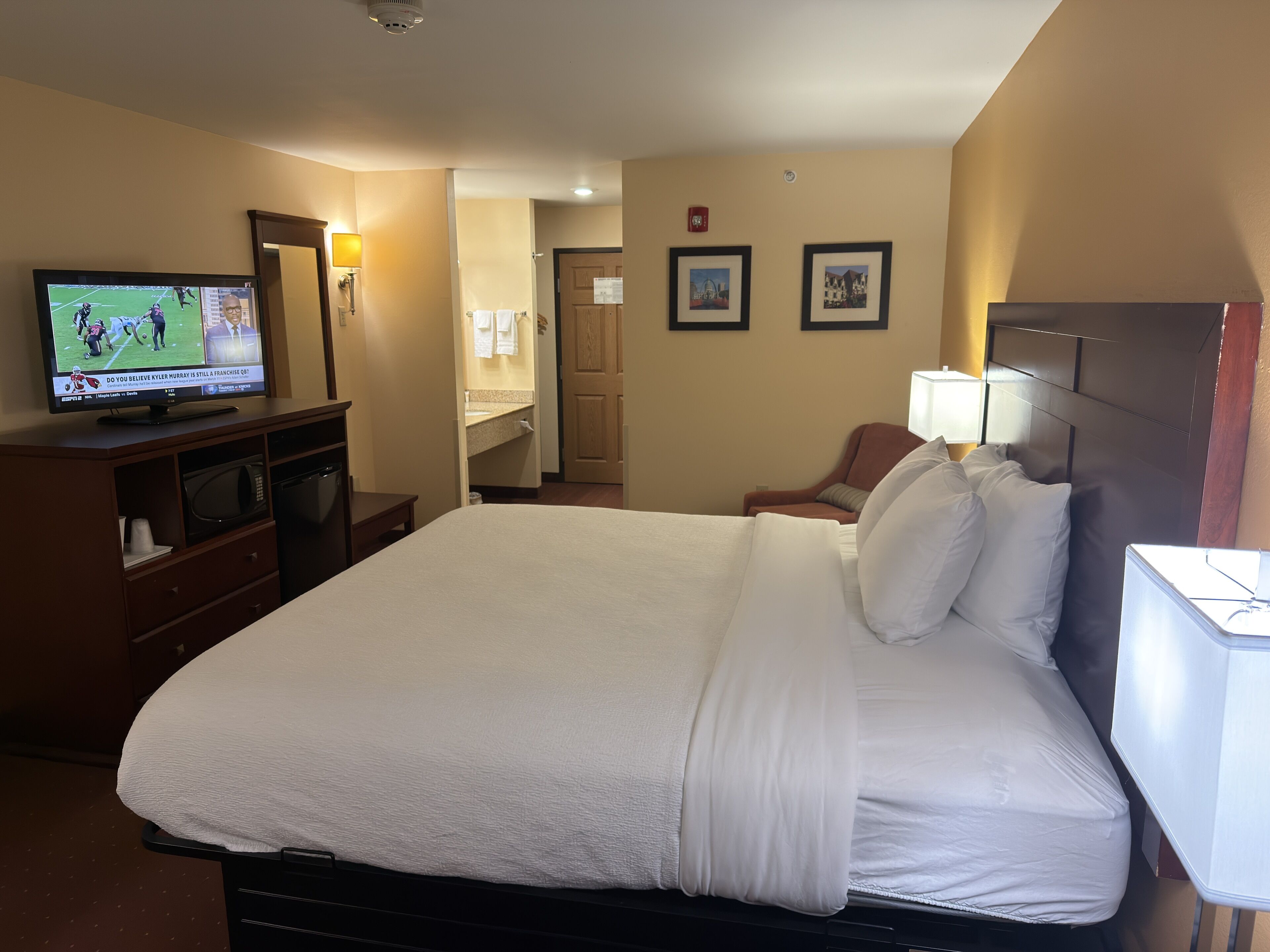 Habitación de hotel con una cama grande, un televisor en una cómoda, una silla, un escritorio y un baño visible a través de una puerta abierta.