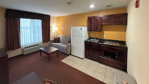A compact hotel room with a kitchenette, a sofa, a small table, and a window with curtains.