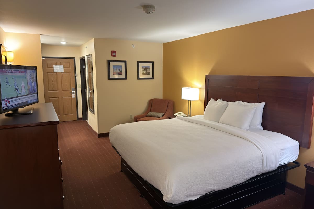 A hotel room with a large bed, a desk with a television, a chair, and a nightstand with a lamp.