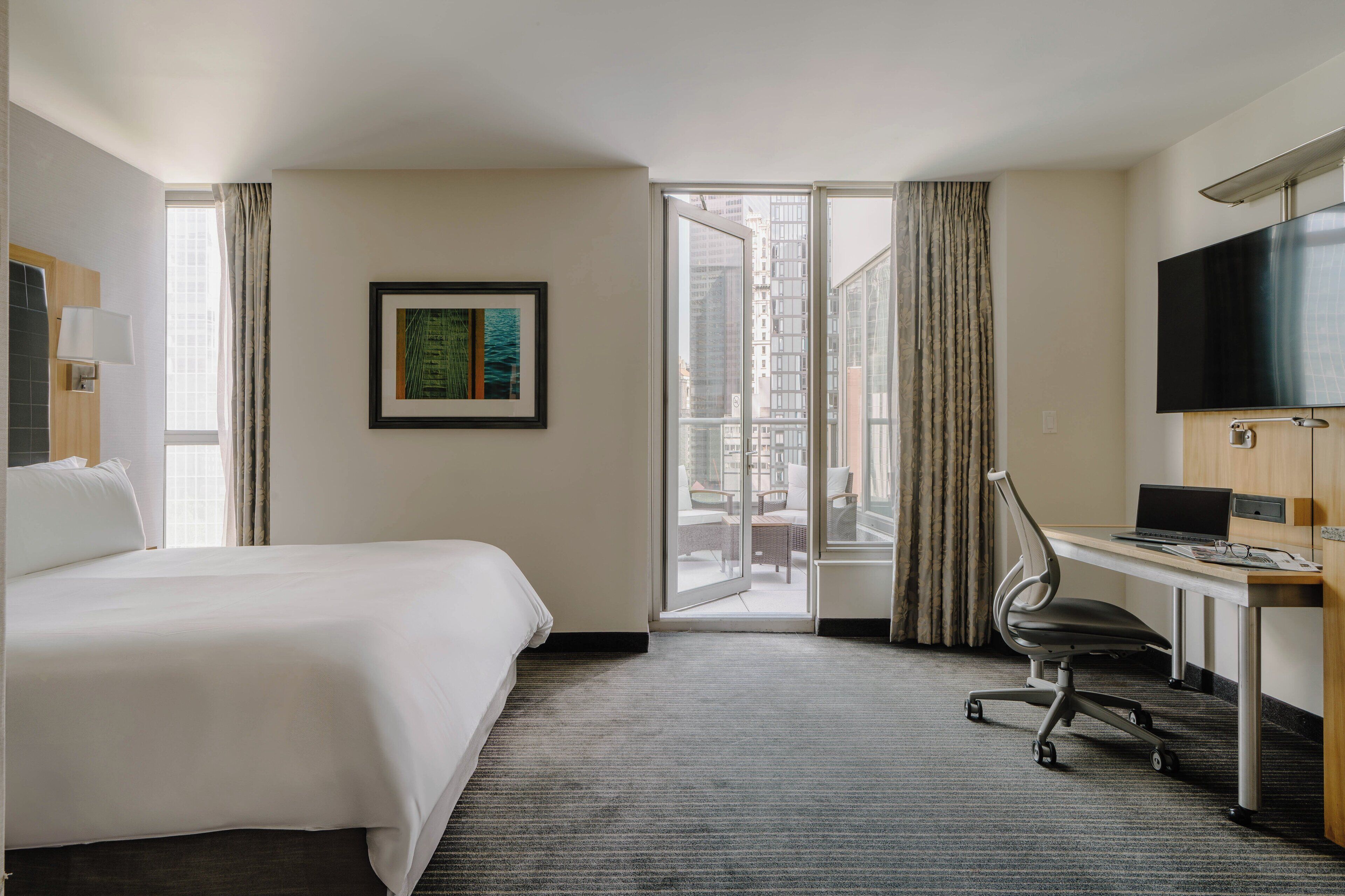 A hotel room with a bed, desk, chair, TV, and a view of the city through a glass door.