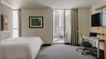 A hotel room with a bed, desk, chair, TV, and a view of the city through a glass door.