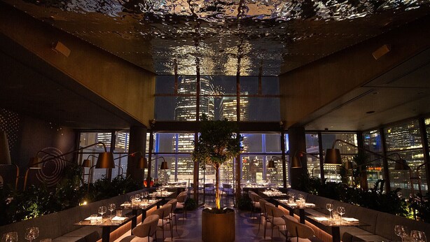 Un restaurant moderne avec un plafond haut, de grandes fenêtres et une plante au centre. Les tables sont dressées avec des verres à vin et des assiettes.