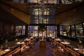A modern restaurant with a high ceiling, large windows, and a central plant. Tables are set with wine glasses and plates. - Club Quarters Hotel World Trade Center, New York (New York)