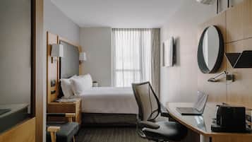 A hotel room with a bed, a desk with a laptop, a chair, a desk lamp, a mirror, and a window with curtains.
