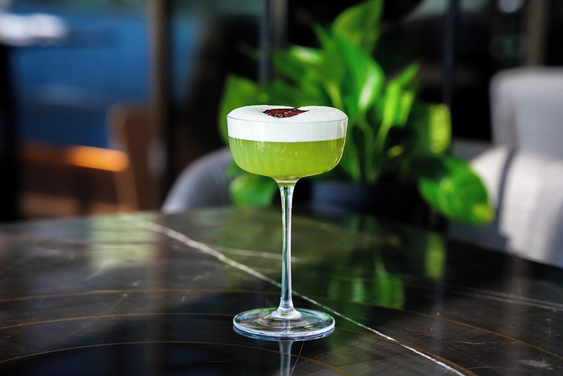 A cocktail in a stemmed glass on a reflective surface with a blurred background of greenery and a chair.