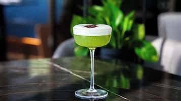 A cocktail in a stemmed glass on a reflective surface with a blurred background of greenery and a chair.