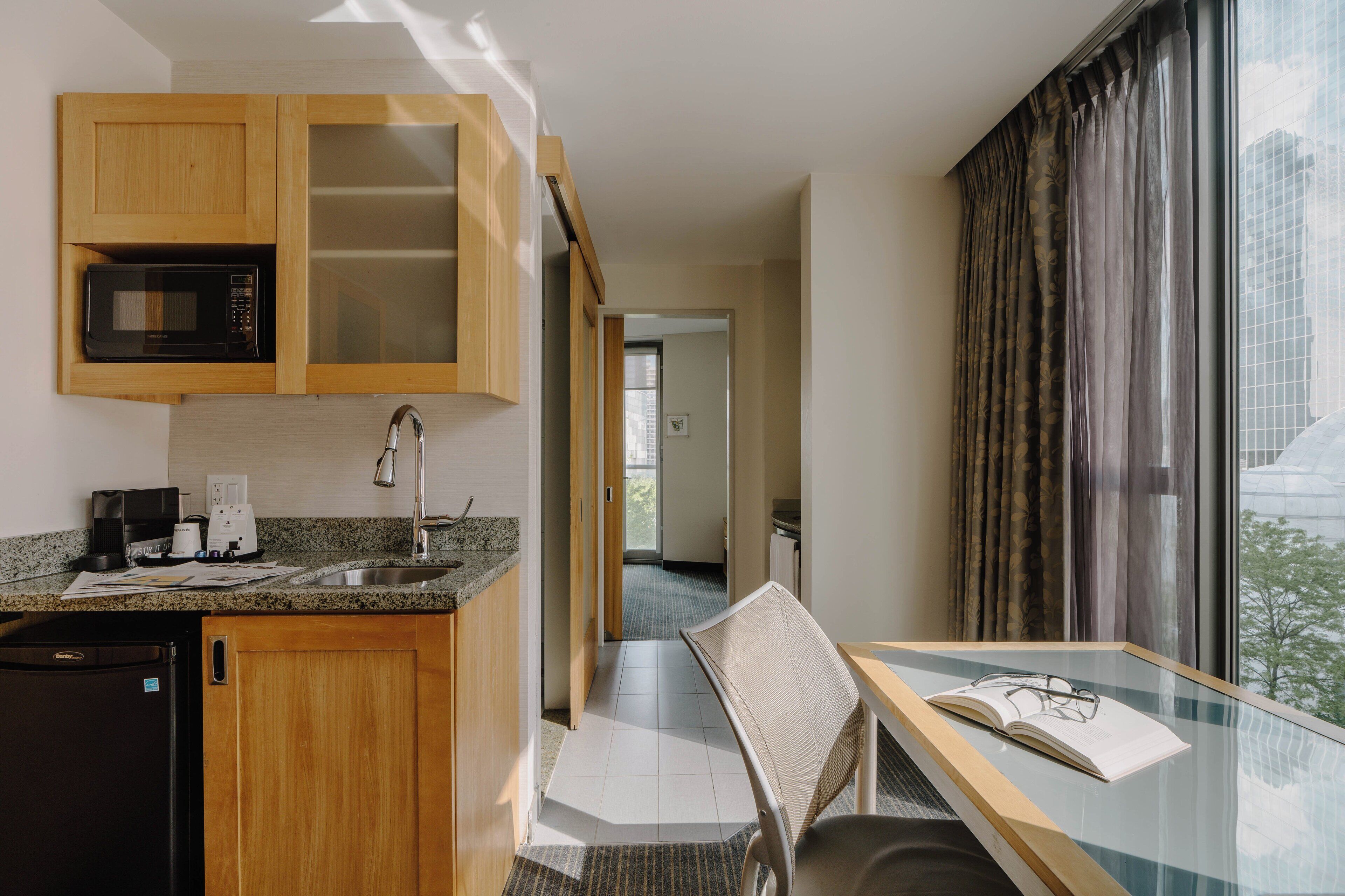 A compact kitchen with a microwave, coffee maker, and sink. There's a dining area with a table and chair, and a window with curtains.