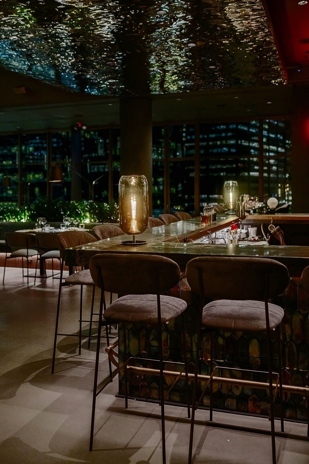 An indoor dining area with a long table, multiple chairs, and a city view through large windows.