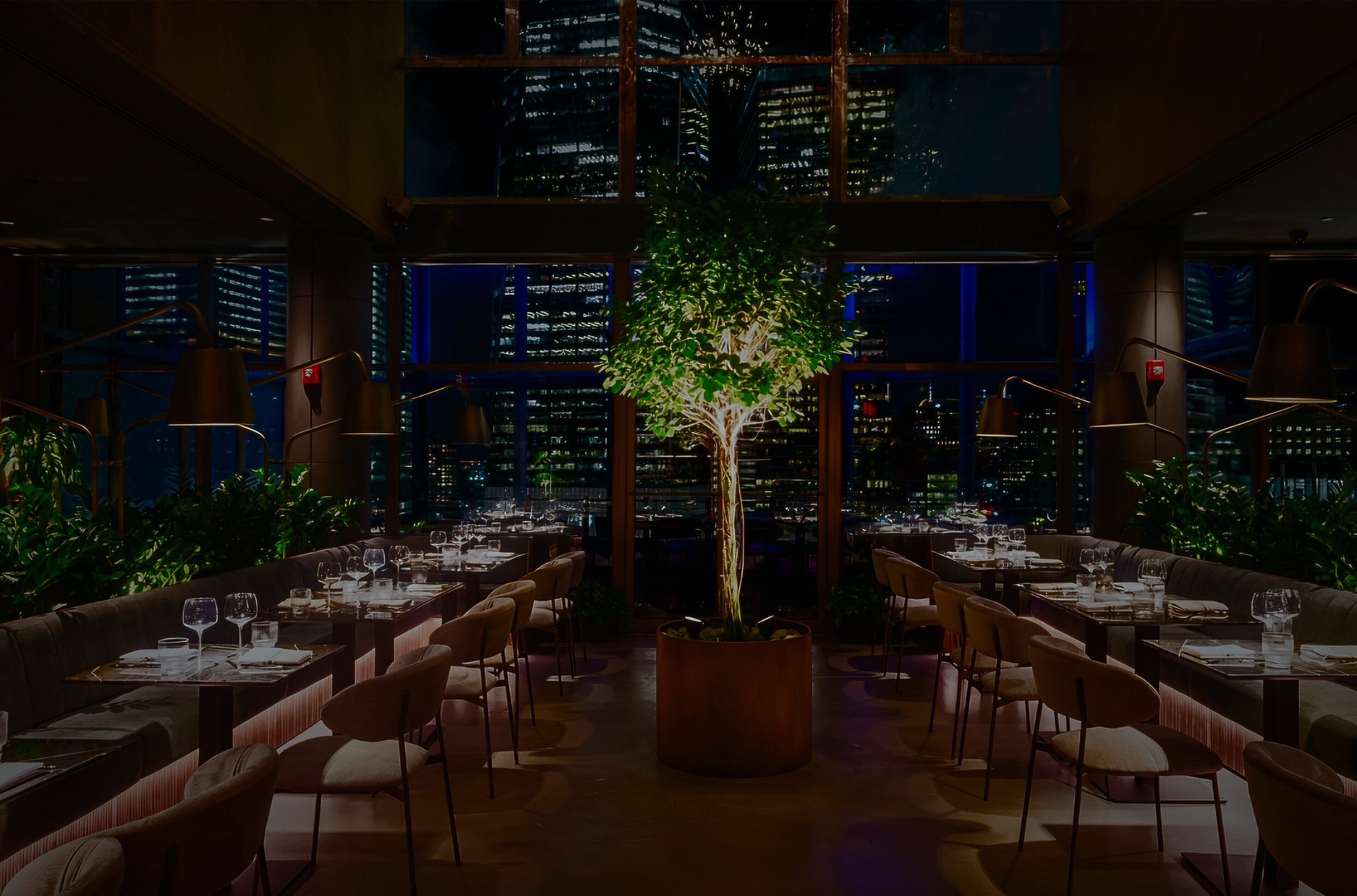 A restaurant with tables set for dining, a central tree, and a cityscape view through large windows.