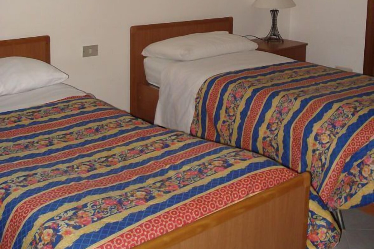 Une chambre avec deux lits simples, chacun agrémenté d’une couvre-lit à motifs et d’une tête de lit en bois. Une table de chevet avec une lampe se trouve entre les deux lits. Deux tableaux sont accrochés au mur, au-dessus des lits.