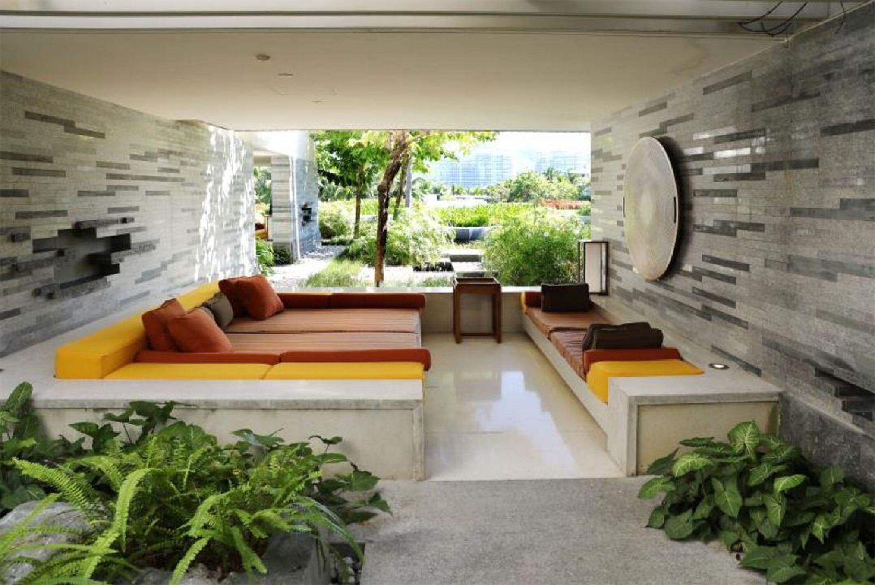 A modern living room with a sofa set, a coffee table, and a TV. There is a view of greenery and a cityscape through the windows.