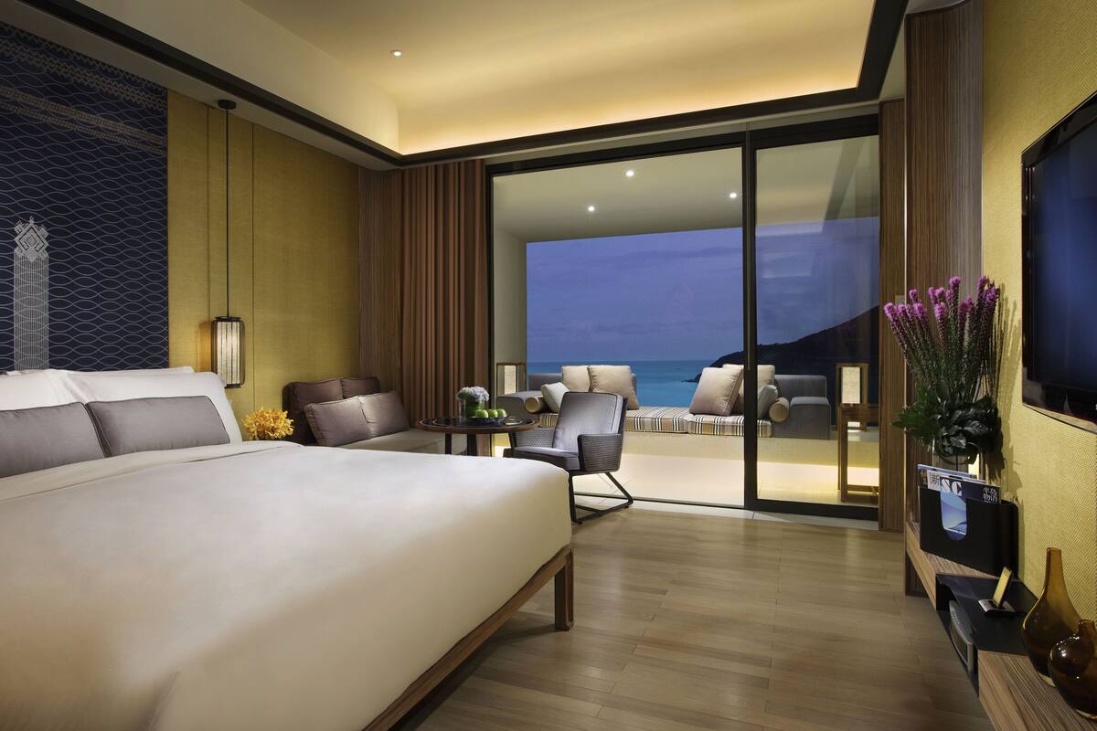 A modern hotel room with a large bed, a seating area with a view of the sea, and a television on the wall.