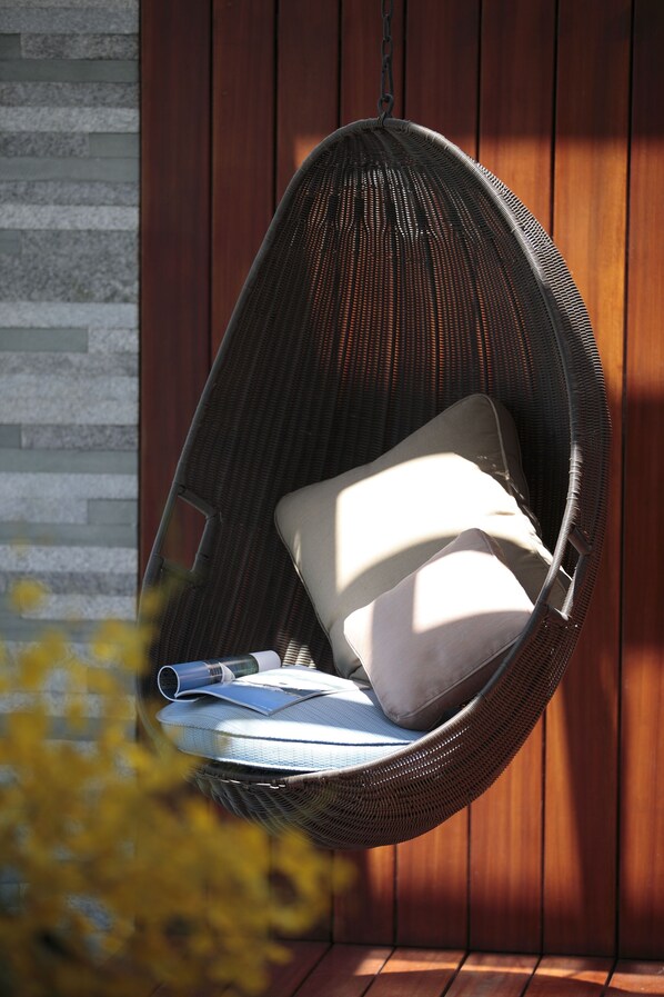 Una silla colgante de mimbre con cojines, una toalla enrollada y un libro, frente a un panel de pared de madera y piedra.