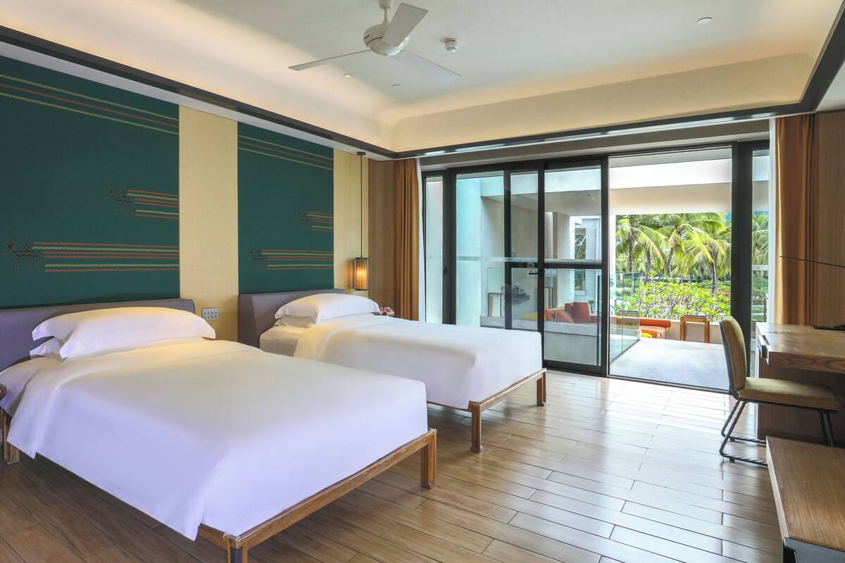 A hotel room with two beds, a desk, and a balcony with a view of palm trees.