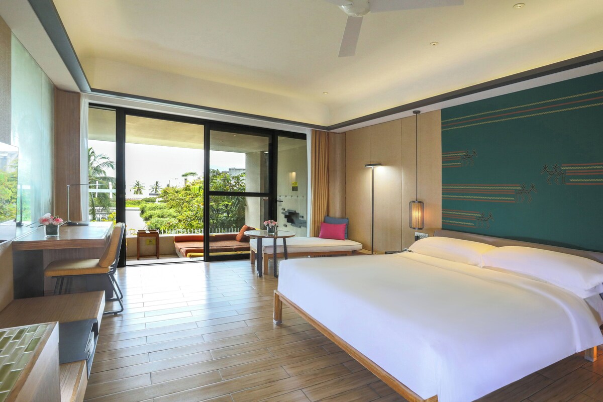 Habitación de hotel con una cama grande, un escritorio con una silla, una mesita con una lámpara y vistas al exterior a través de una puerta corrediza.