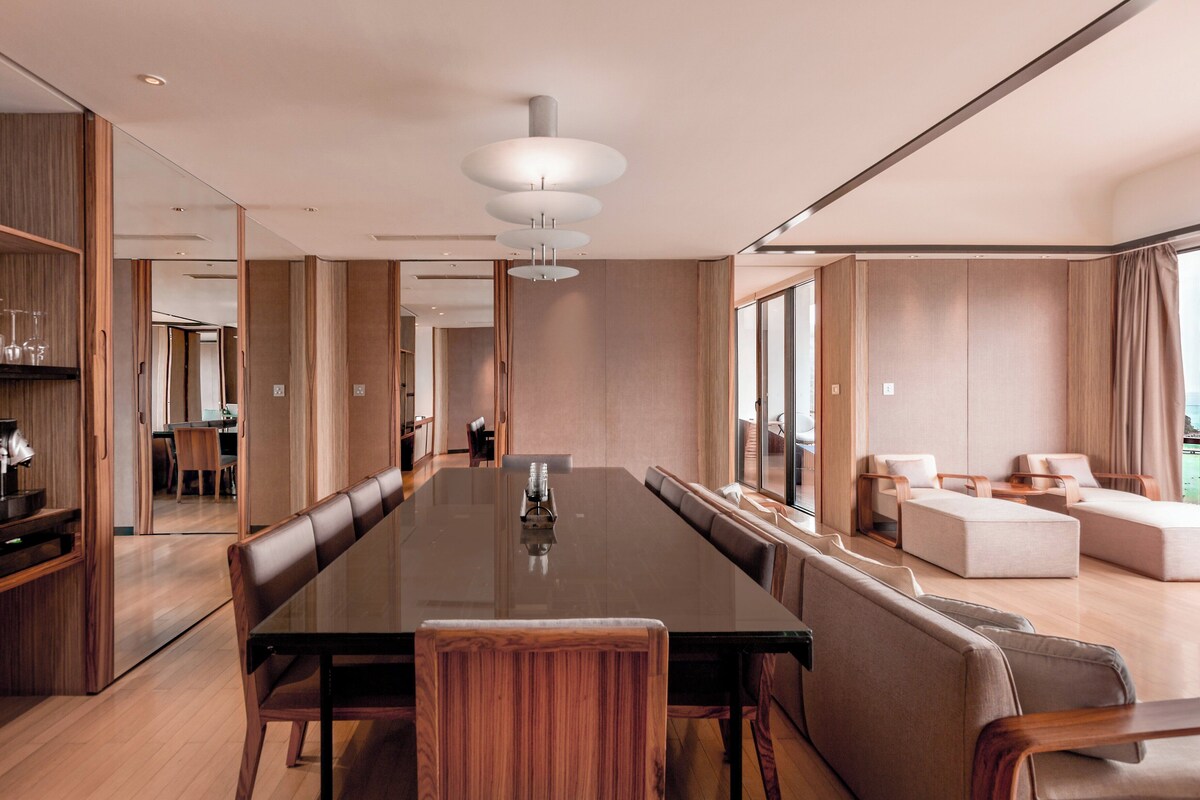 Una habitación de hotel moderna con una amplia mesa de comedor, asientos cómodos, una zona de cocina y ventanas grandes con vistas.