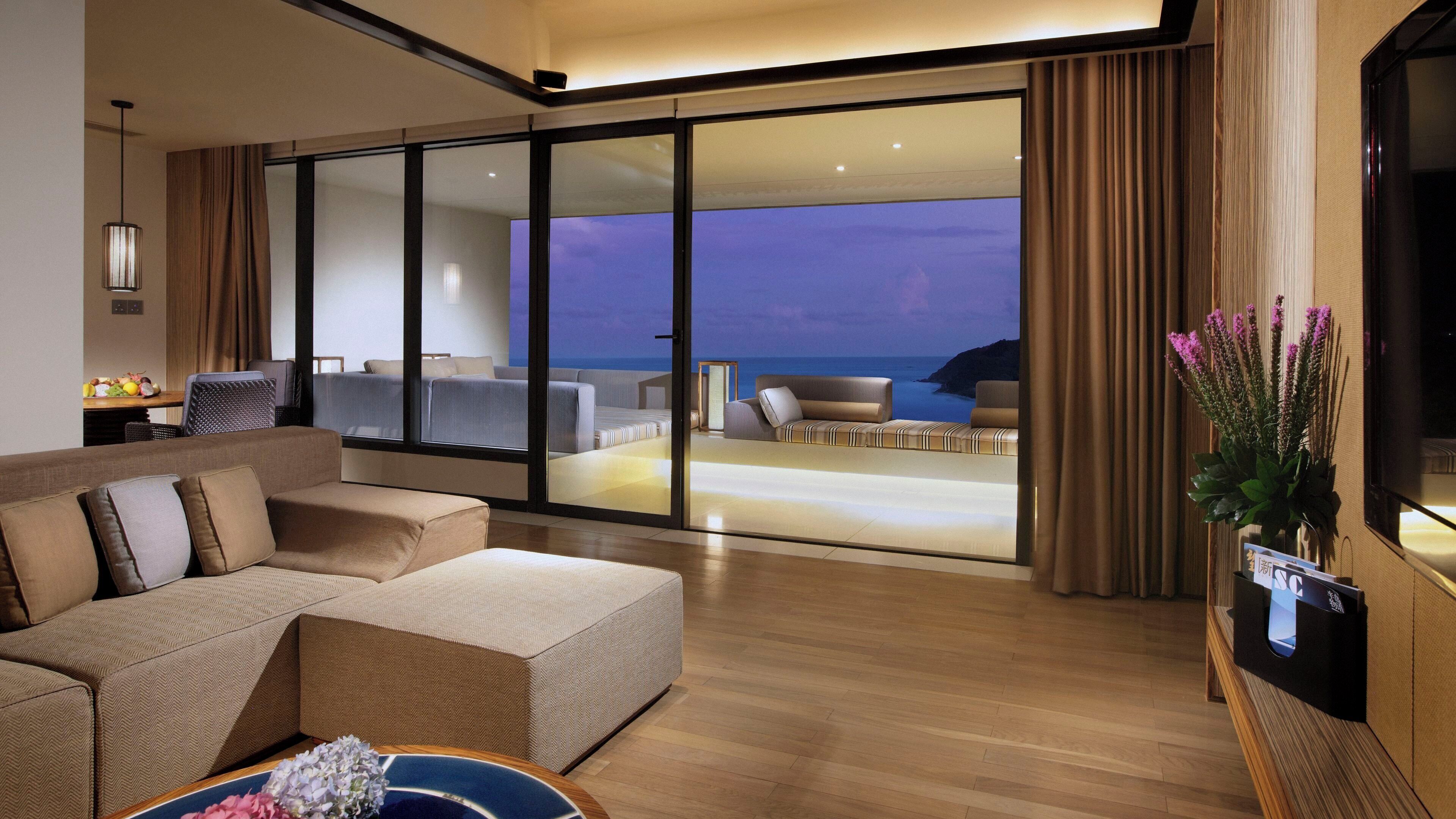 A modern hotel room with a large sofa, a dining area, and a balcony with a view of the sea.