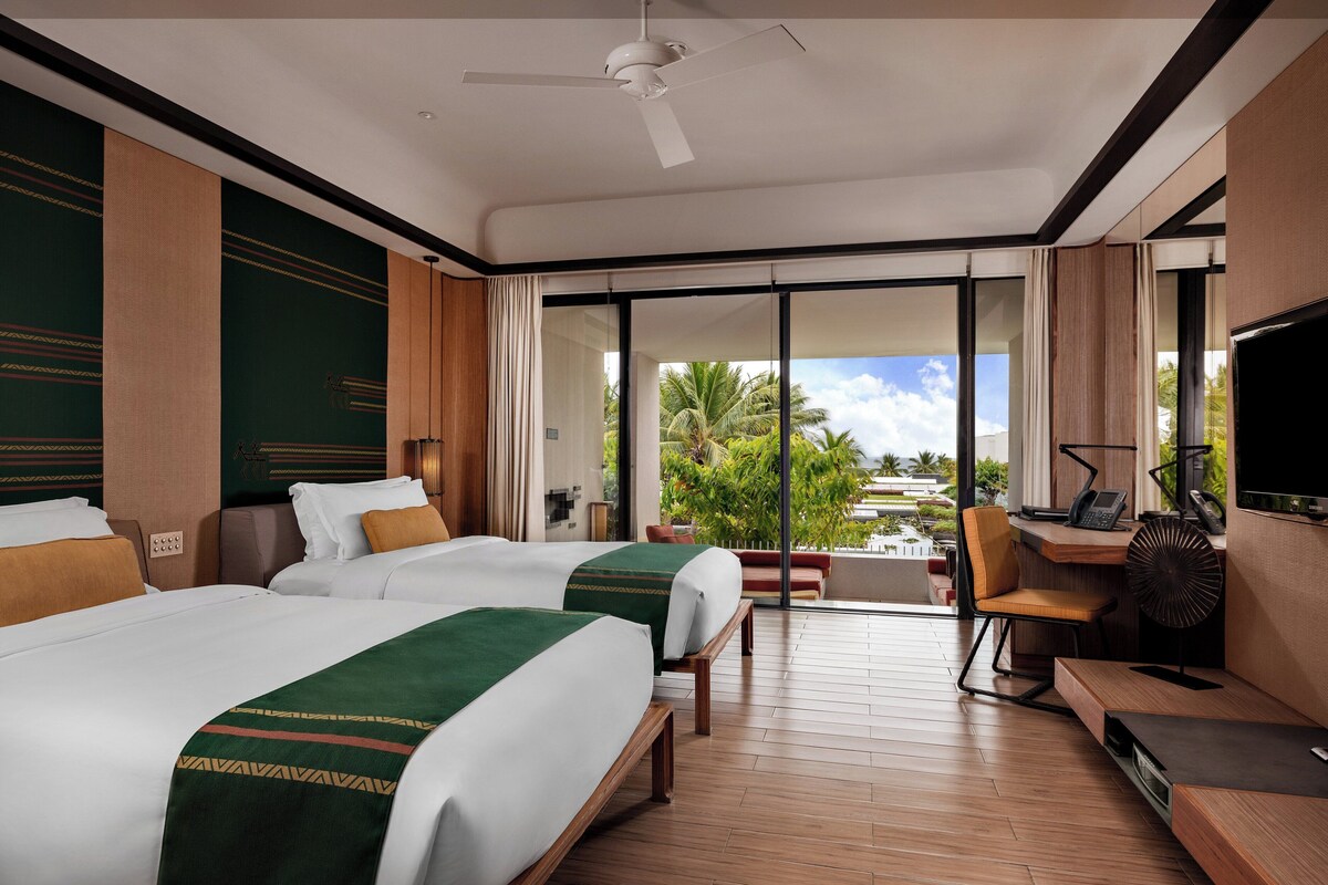 A hotel room with two beds, a desk, a chair, a TV, and a view of the outdoors.