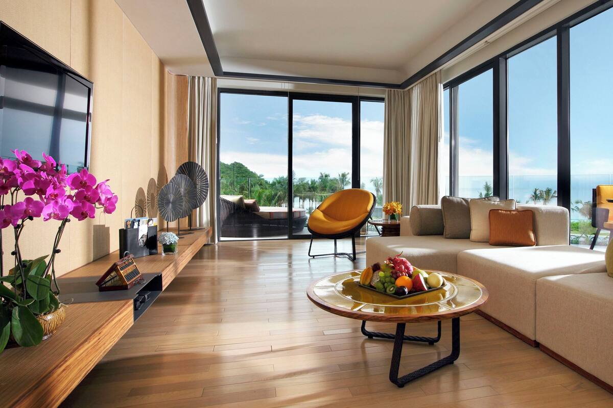 A modern living room with a large sofa, a coffee table with fruit, and a view of the ocean.