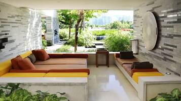 A modern living room with a sectional sofa, a coffee table, and a TV. There is a view of a garden and buildings outside.