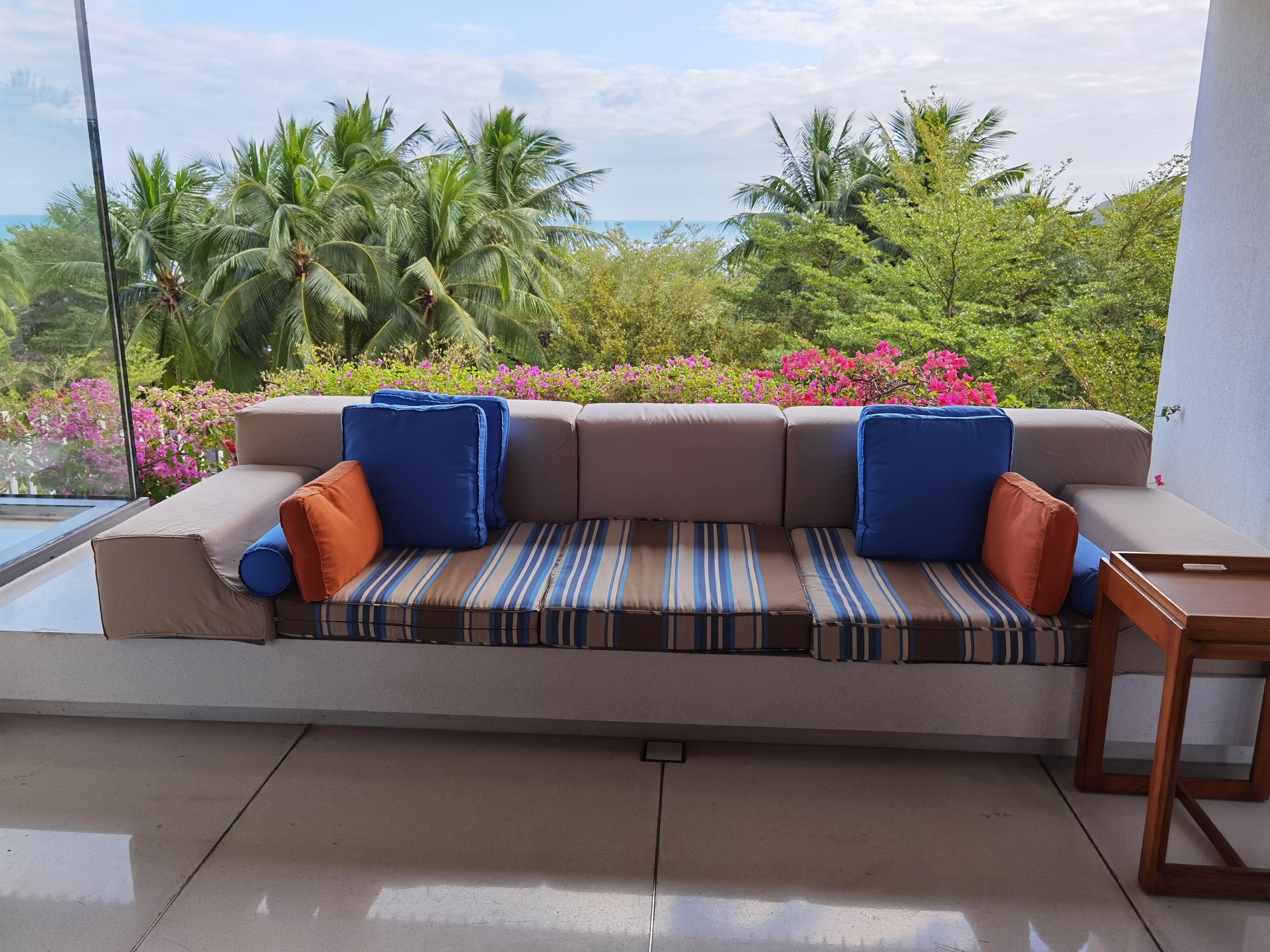 A sofa with striped cushions, a wooden side table, and a view of lush greenery and pink flowers.