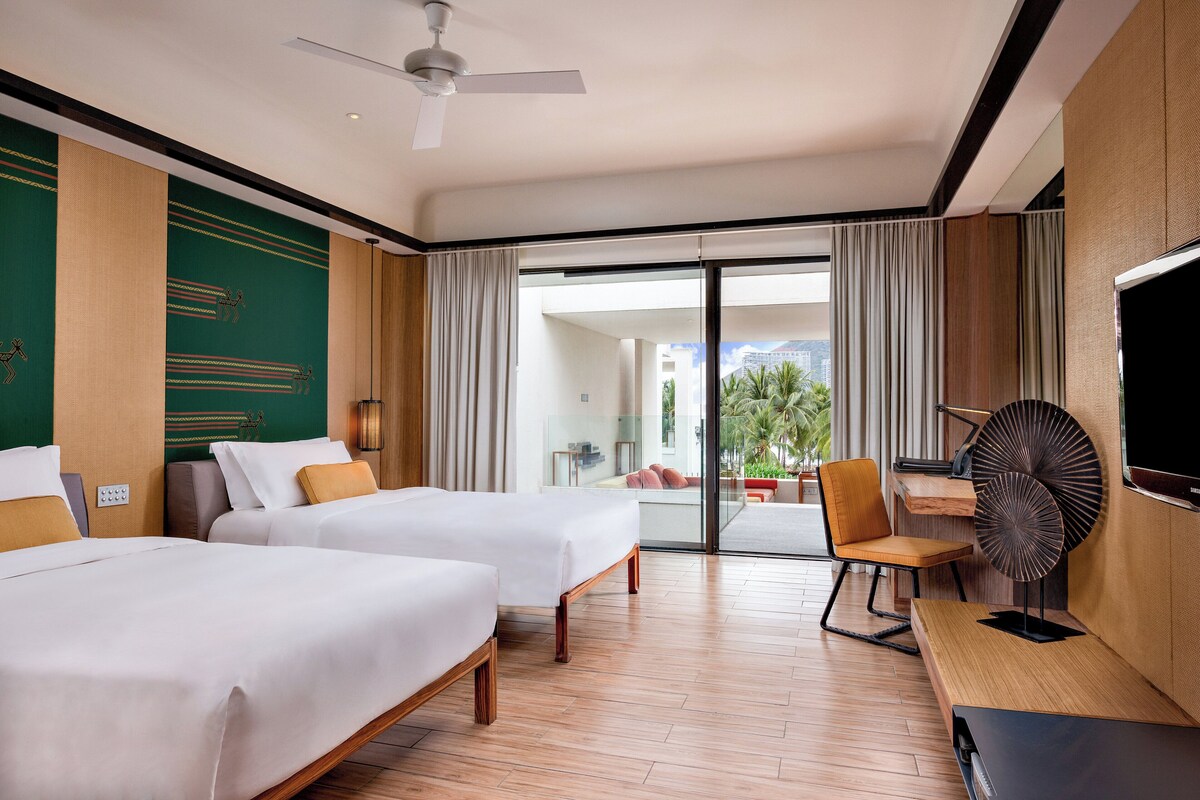 A hotel room with two beds, a desk with a chair, a TV, and a large window with curtains.