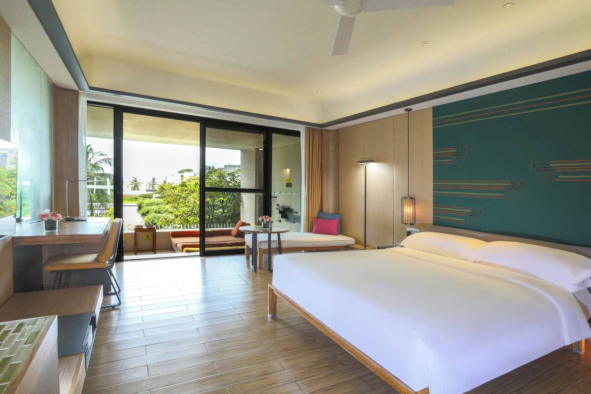 A hotel room with a large bed, a desk with a chair, a small table with a lamp, and a view of the outdoors through a sliding door.