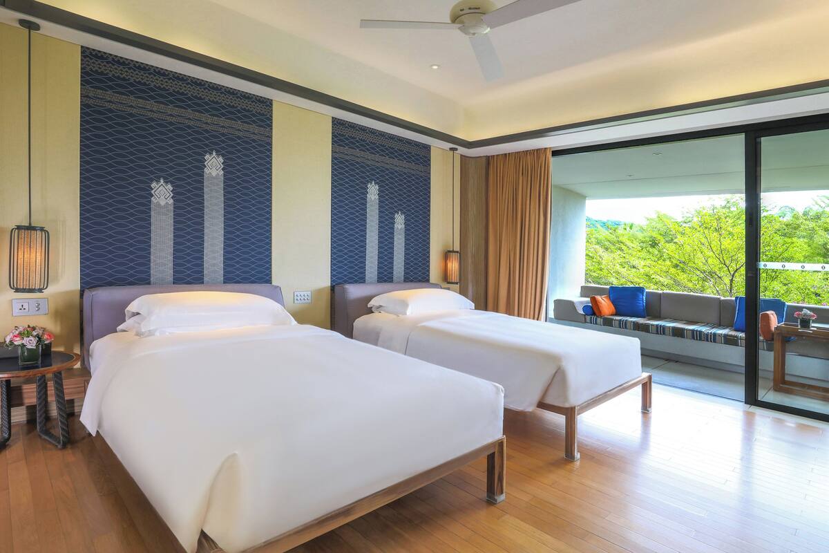 A hotel room with two beds, a sofa, a dining table, and a large window with a view of greenery.