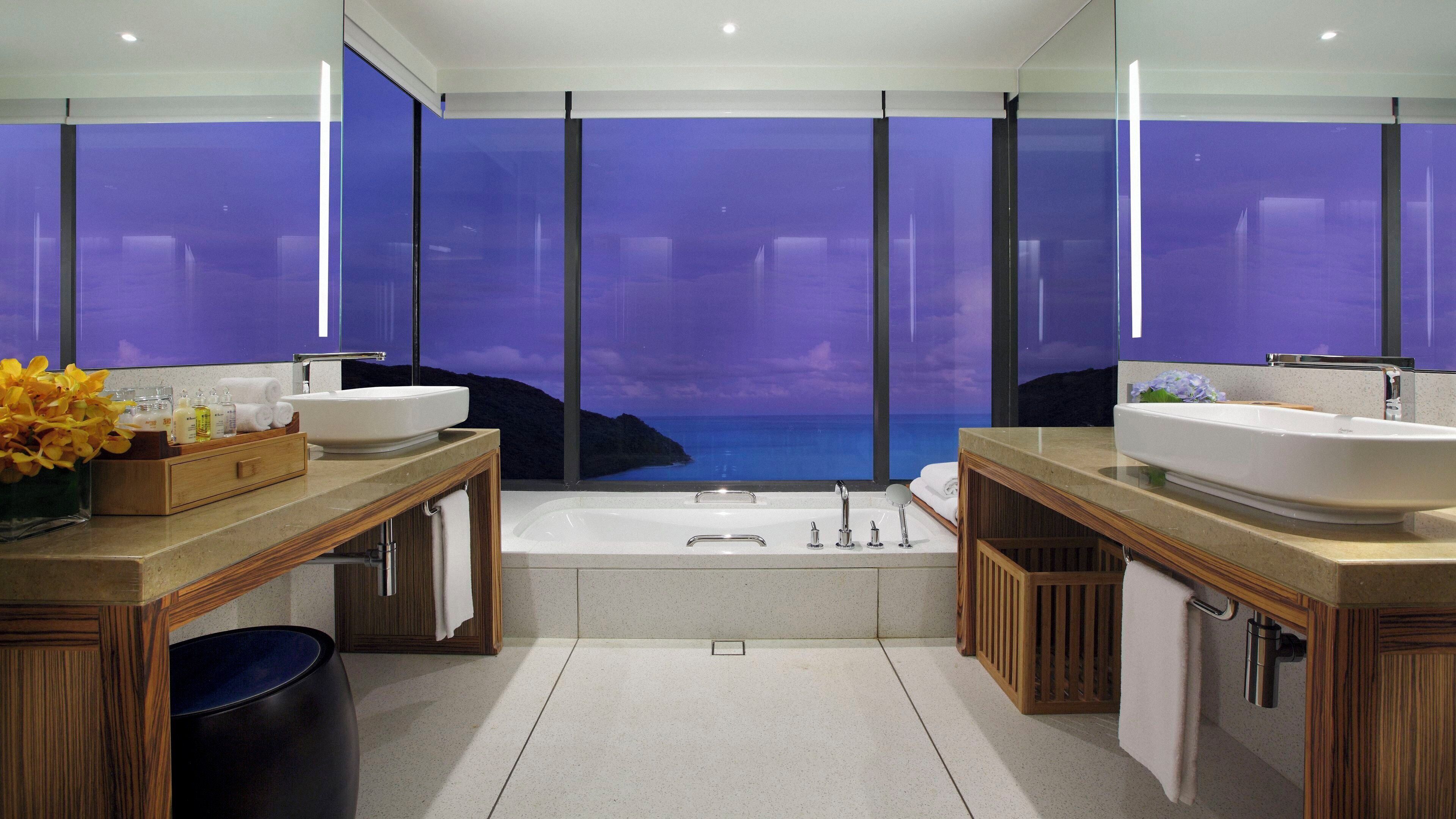A modern bathroom with two sinks, a bathtub, and a large window with a view of the sea.