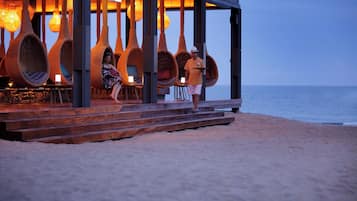 A beachfront establishment with wicker chairs and tables, a wooden deck, and a view of the sea.